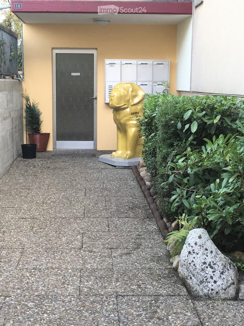 A building entrance with a door, a golden lion statue, and a small garden with plants and a rock.