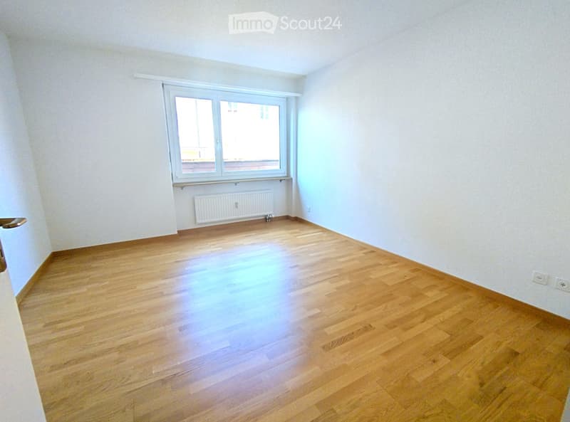 Empty room with wooden flooring, white walls, and a large window with white frame. A radiator is visible below the window.