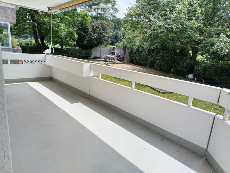 A modern, spacious balcony with white railings and a concrete floor, overlooking a lush green garden with trees and a small seating area.