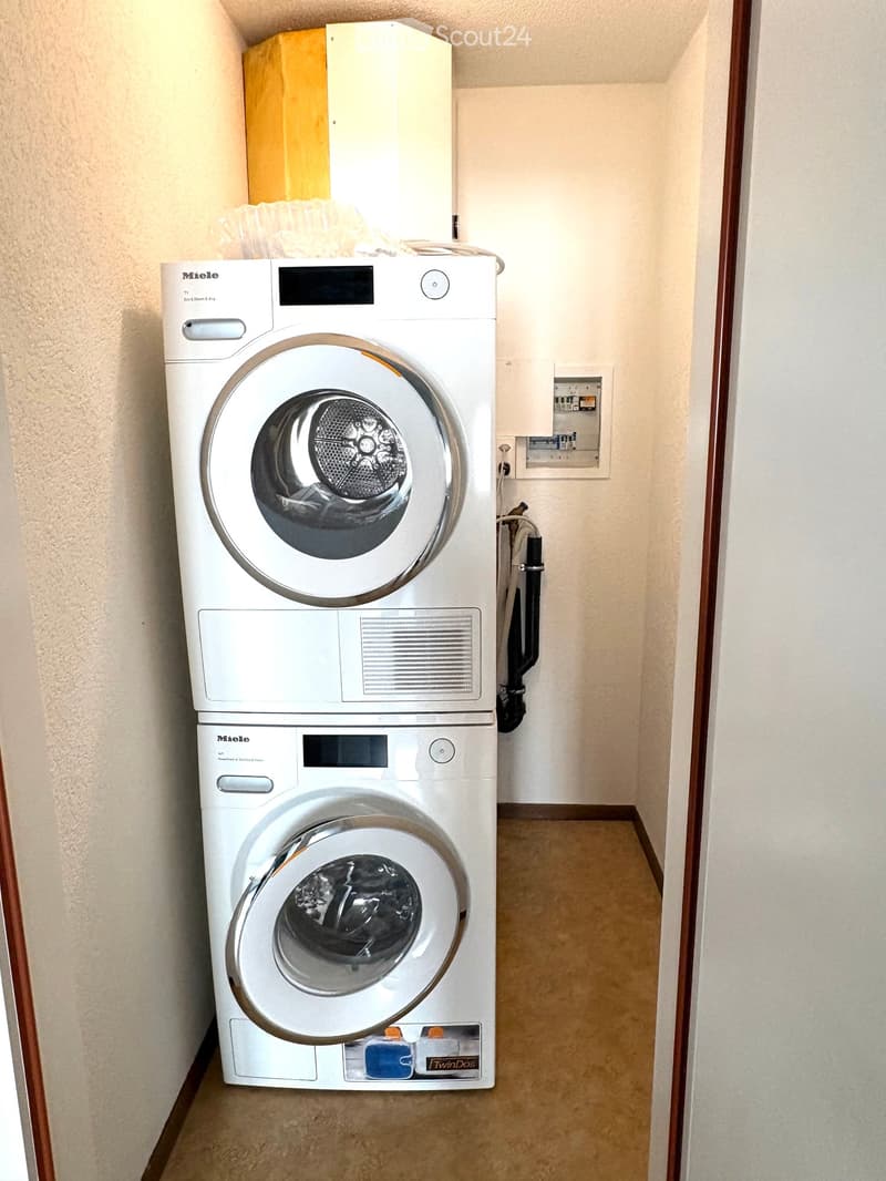 A narrow hallway with beige walls and carpeted floor, featuring two stacked Miele washing machines with control panels and a small electrical panel on the wall.