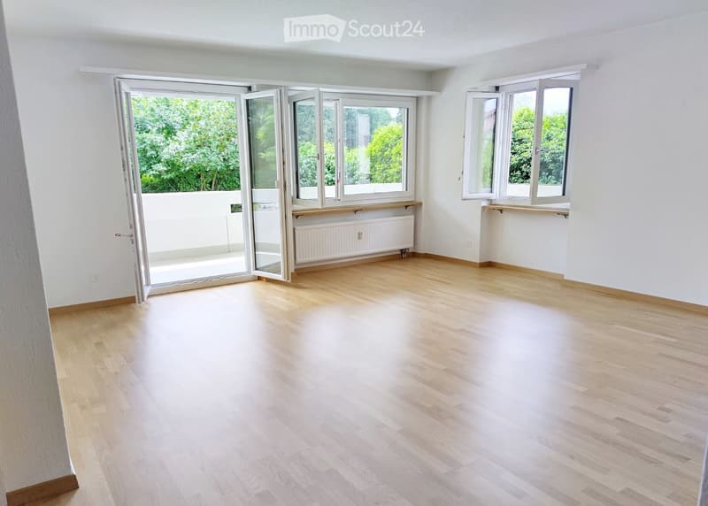 Empty room with light wooden flooring, white walls, large windows, and a sliding glass door leading to a balcony.