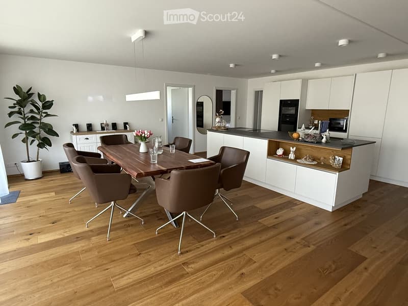 Furnished Room. Modern dining area with wooden table, brown chairs, and a white cabinet. A plant and flowers add a touch of nature. The kitchen has a counter, microwave, and oven.