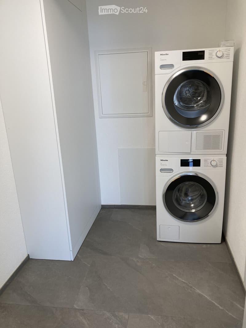 Bathroom. A small, modern bathroom with a white washing machine and dryer stacked vertically, grey tiled floor, and white walls.