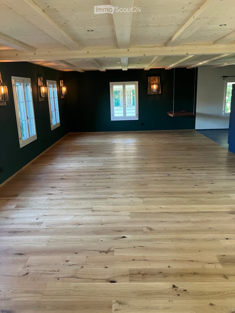 Empty room with wooden floor, dark green walls, exposed wooden ceiling beams, and several windows with white frames.