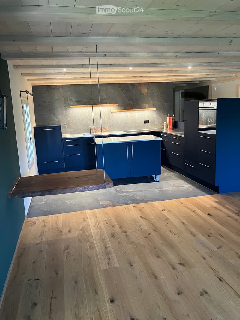 Modern kitchen with dark blue cabinets, stainless steel appliances, and a wooden island. Exposed wooden beams on the ceiling and a large wooden floor.