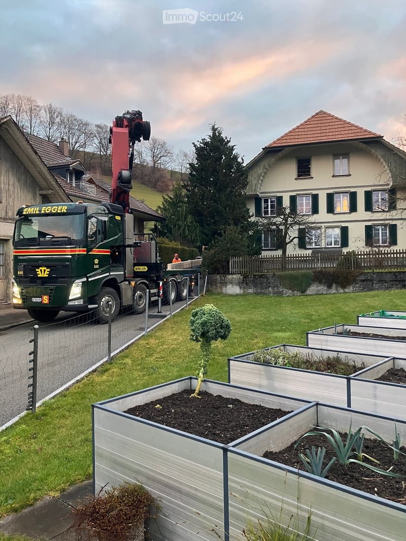 A garden area with raised beds, a truck with a crane, and a house in the background.