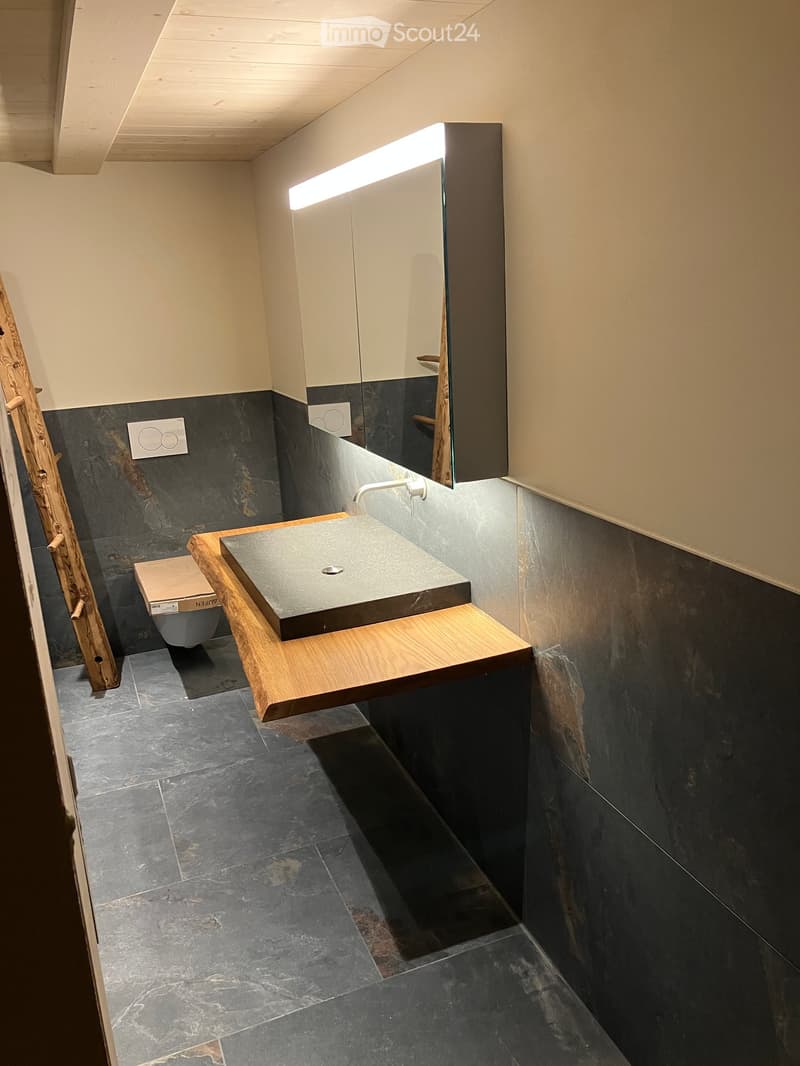 Modern bathroom with a wooden sink console, stone sink, dark tiled walls and floor, and a large mirror with built-in lighting.