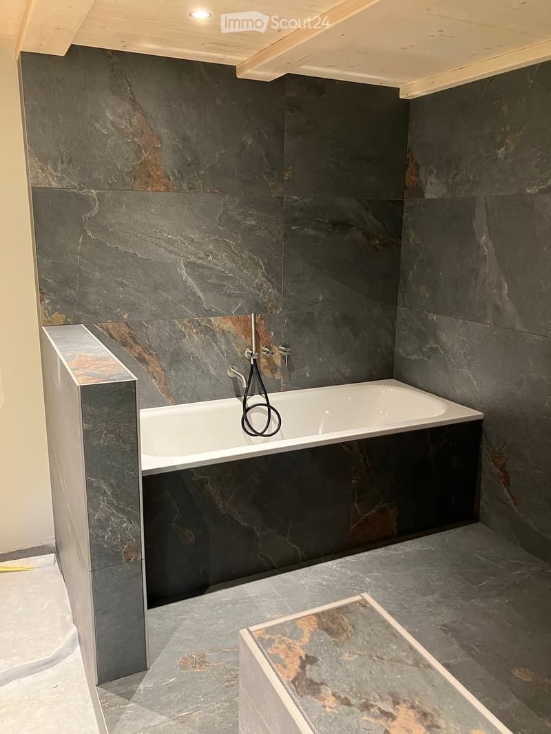 Modern bathroom with dark marble walls and floor, white bathtub, sleek faucet, and wooden ceiling.