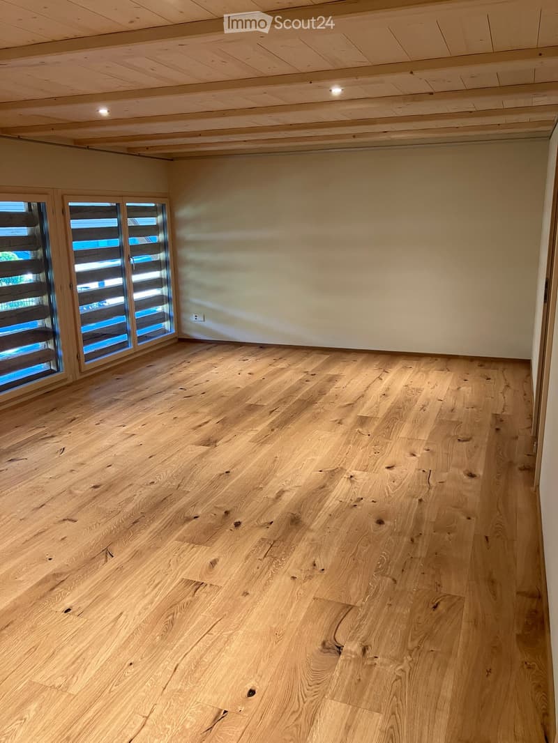 Empty room with light wooden flooring, white walls, and a large window with blinds allowing natural light to enter.