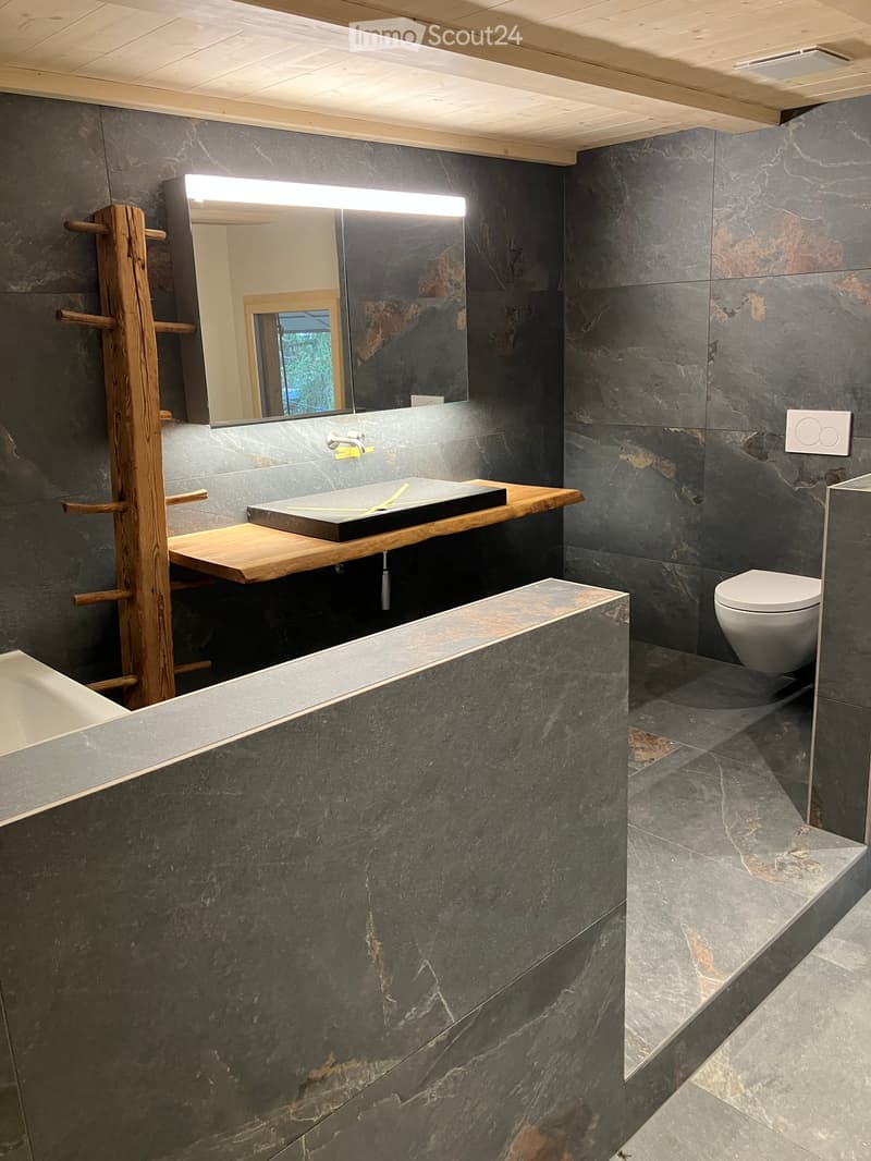 Modern bathroom with dark grey marble tiles, a wooden sink console, a toilet, and a large mirror with built-in lighting.