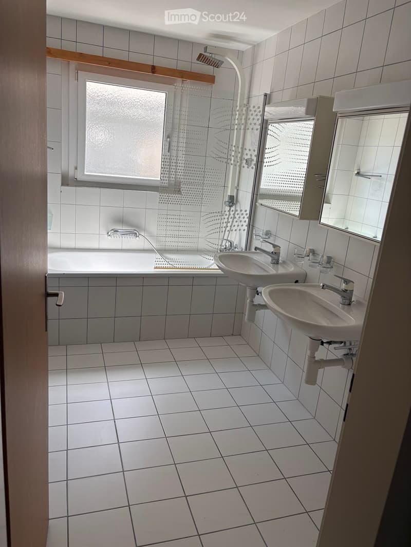 Modern bathroom with white tiled walls and floor, featuring a bathtub, two sinks, a shower area with glass enclosure, and a mirror above one sink.