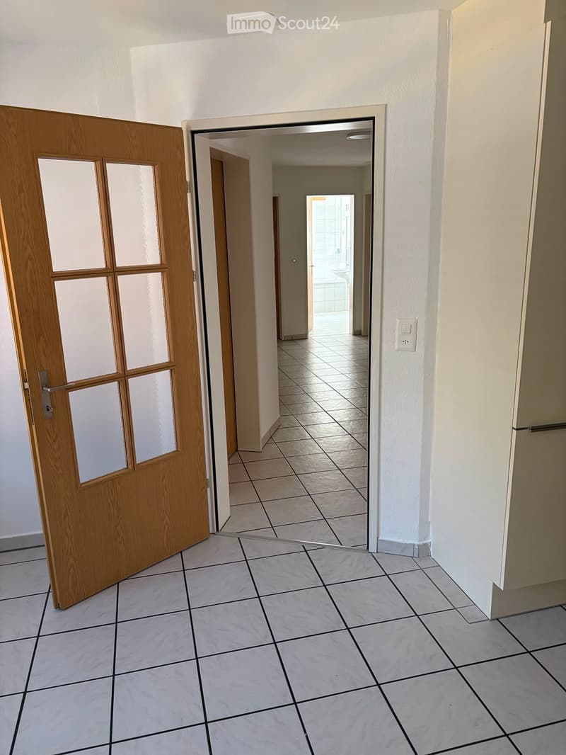 Empty hallway with tiled flooring, white walls, and a wooden door with glass panels on the left leading to another room.