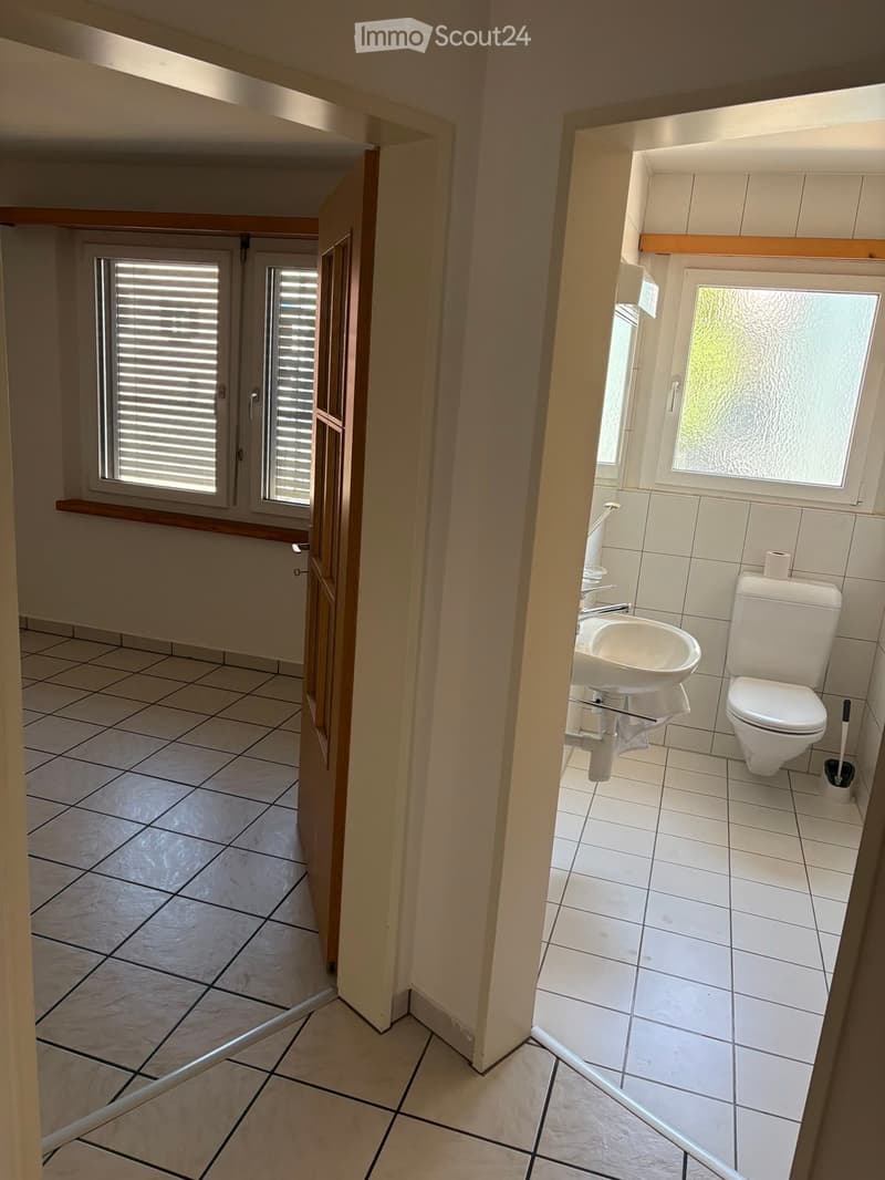 Modern bathroom with white tiles, a sink, toilet, and a window with frosted glass. The floor is tiled, and the walls are painted in a light color.