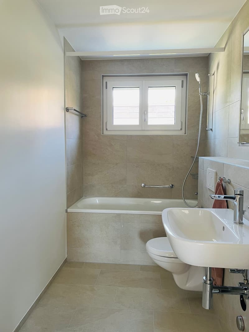 Modern bathroom with beige tiles, bathtub, toilet, sink, and shower. White fixtures and a window for natural light.