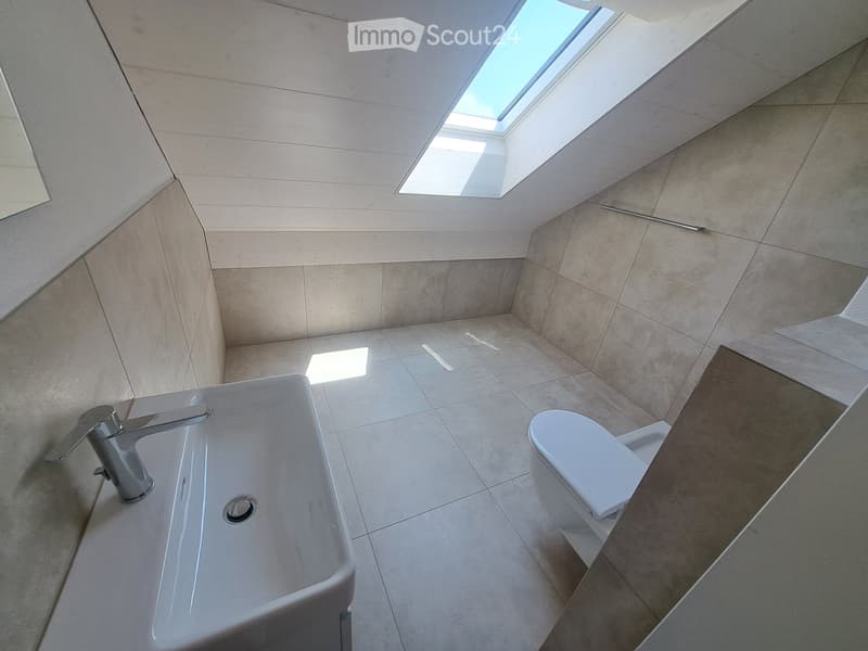Bathroom with white tiles, a sink, a toilet, and a mirror. There is a skylight for natural light.