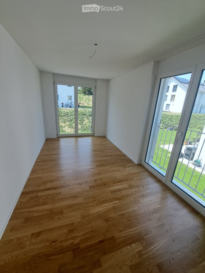 Empty room with white walls, wooden floor, and sliding glass doors leading to a balcony with a view of a grassy area and a house.