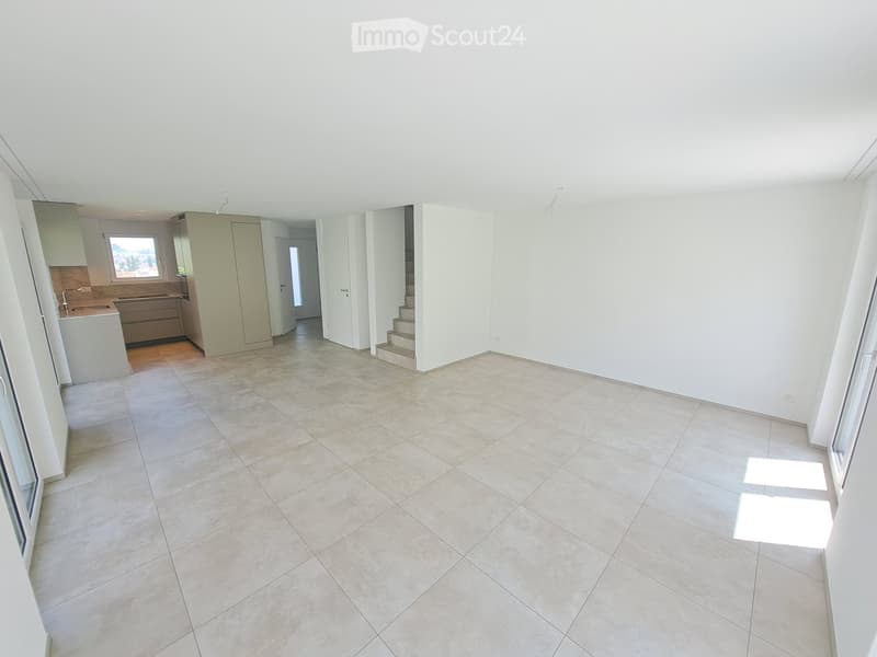 An empty room with white walls, tiled floor, and a staircase leading to the upper floor. There are cabinets and a sink in the kitchen area.