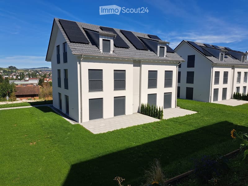 Modern two-story house with white exterior, grey roof, solar panels, multiple windows, and garage doors. The house is surrounded by a well-maintained lawn and garden area.