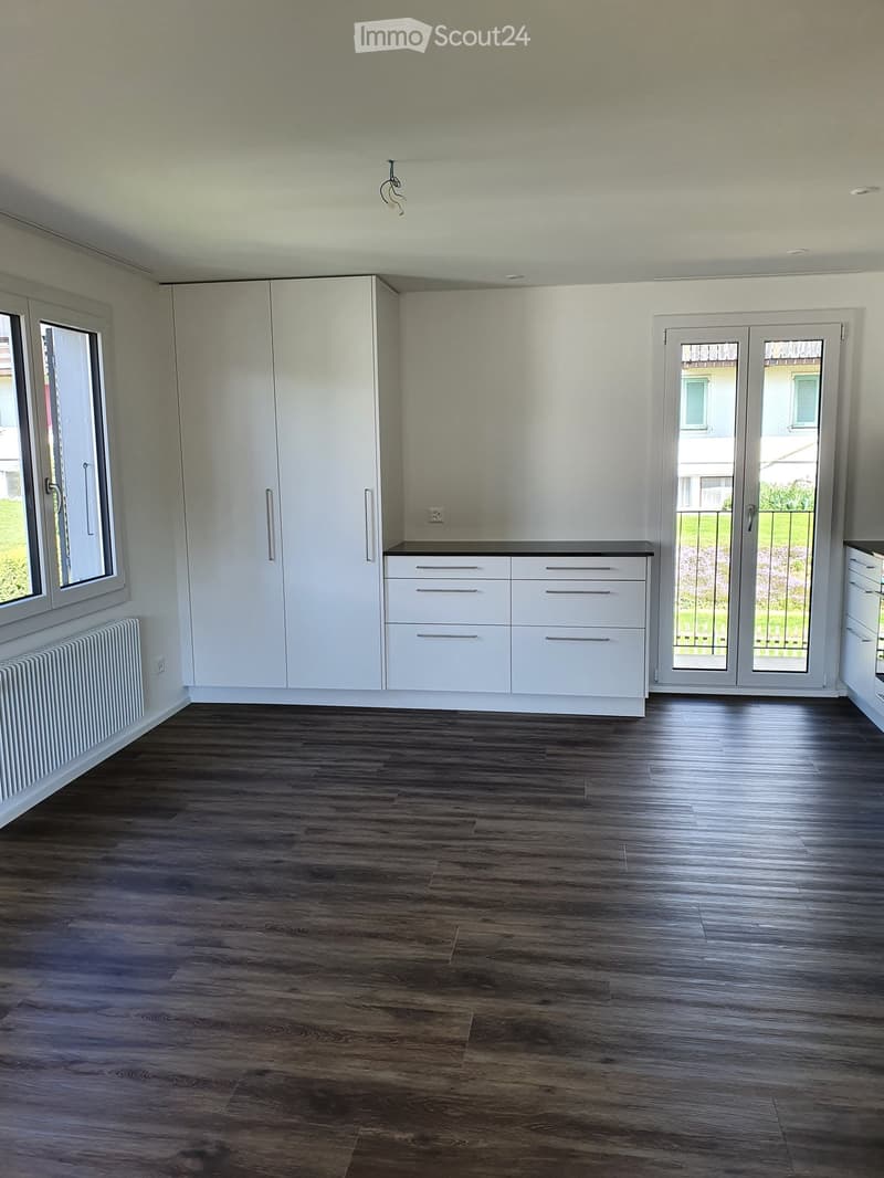 Empty room with white walls, wooden floor, large windows, white cabinets, and a sliding glass door leading to a garden.