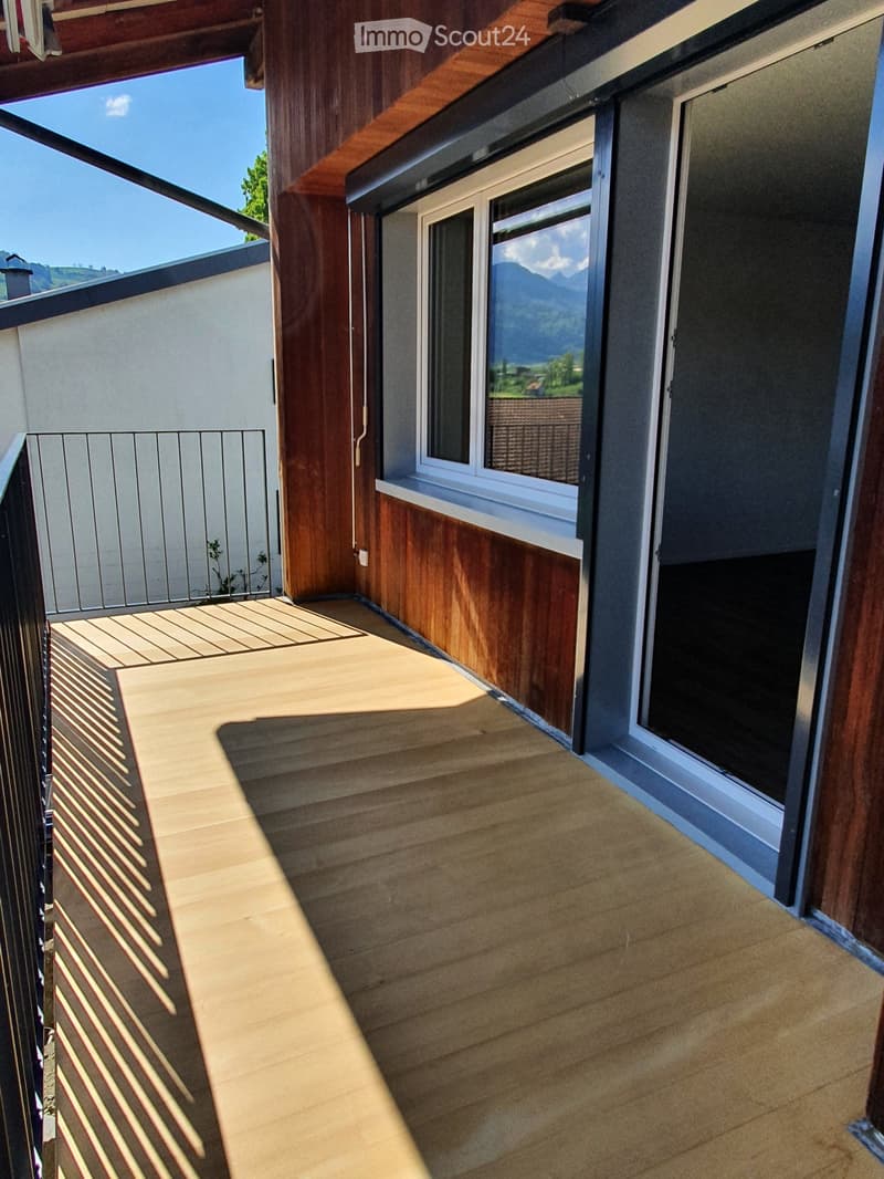 A balcony with wooden flooring and a metal railing. The balcony has a view of the mountains and a clear blue sky. The balcony is attached to a building with a white wall and a window with a white frame.