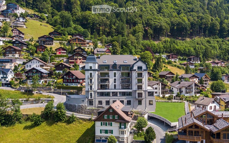 Aerial view of a modern apartment building surrounded by houses and trees, set on a hillside with a scenic valley below.