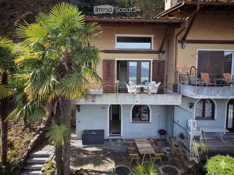 Two-story house with balcony, wooden table and chairs, grill, storage box, and palm trees in the front yard