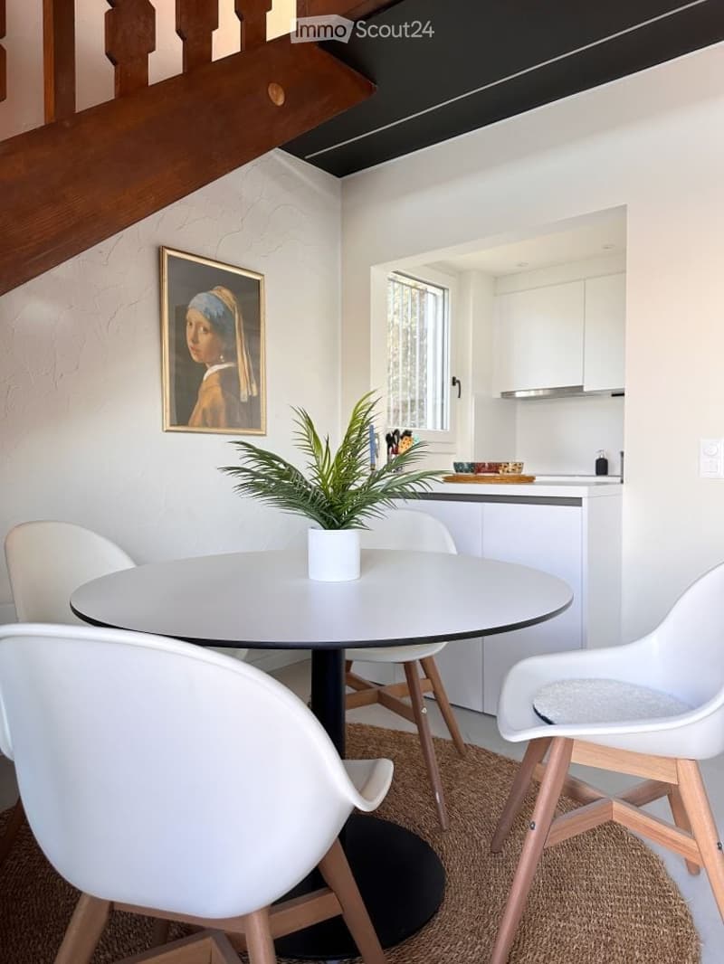 Dining area with a round table, white chairs, a plant, a picture frame, and a small kitchen area