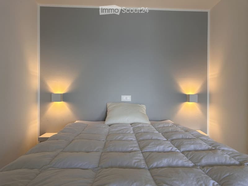 A bedroom with a bed, a white pillow, gray walls, and two wall-mounted lights.