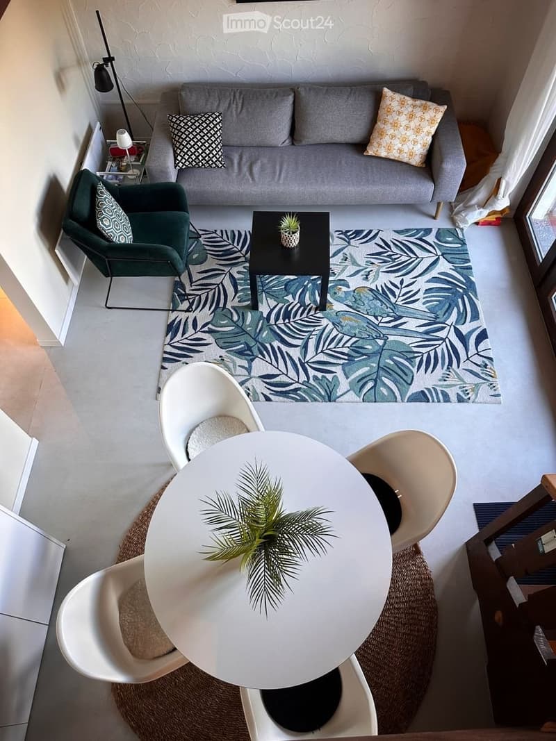Open living area with a gray couch, a green armchair, a round table, and a rug with a tropical pattern.