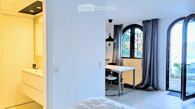 A room with a desk, chair, and a bed. There is a sink and cabinet in the bathroom. There is a window with a view of the outside.