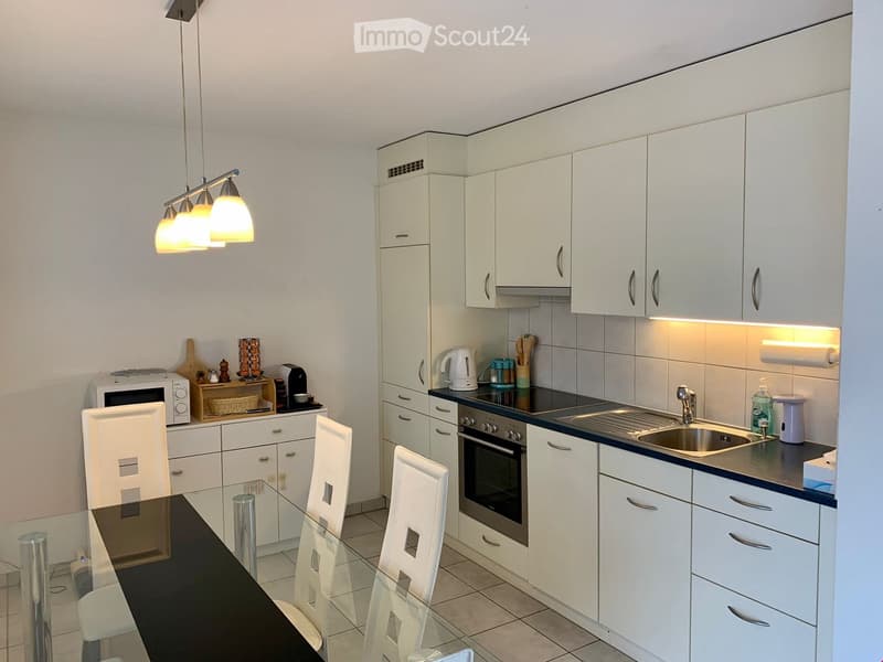 Modern kitchen with white cabinets, black countertops, and a glass dining table. Equipped with a microwave, coffee machine, and a built-in oven.
