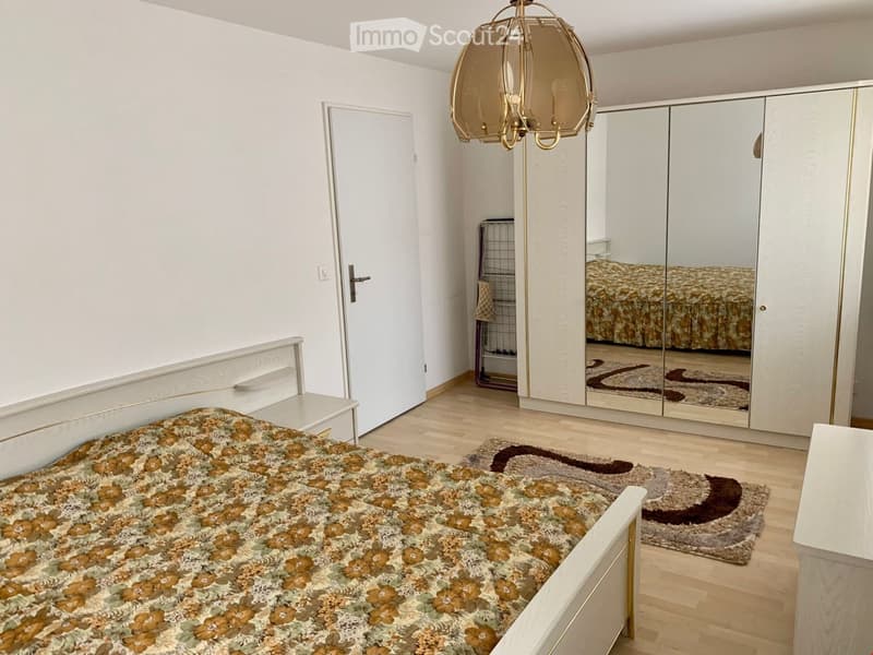 Spacious bedroom with a bed, nightstand, rug, wardrobe, lamp, mirror, and a rack. The room has wooden floors and white walls.