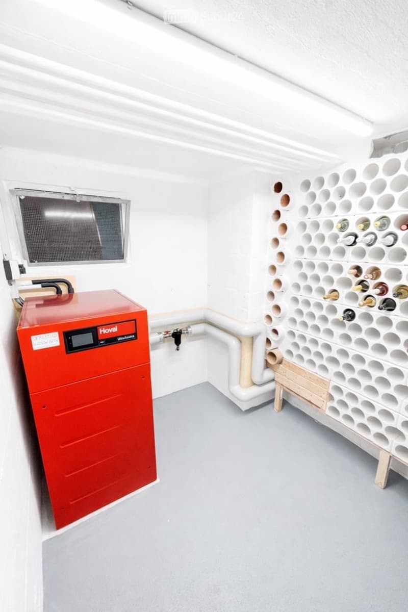 A room with a red heating unit, a white wall with a window, a white ceiling with a light, a white wall with a white pipe, and a white wall with a white pipe.