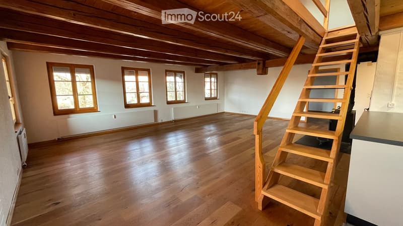 Large room, wooden floors, visible beams, white walls, windows, radiators, stairs leading to upper floor