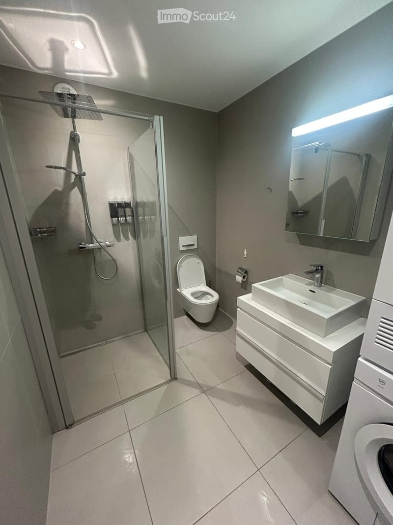Modern bathroom with a glass shower enclosure, white toilet, white sink, and a mirror. The floor and walls are tiled in white. There is a washing machine on the right side.