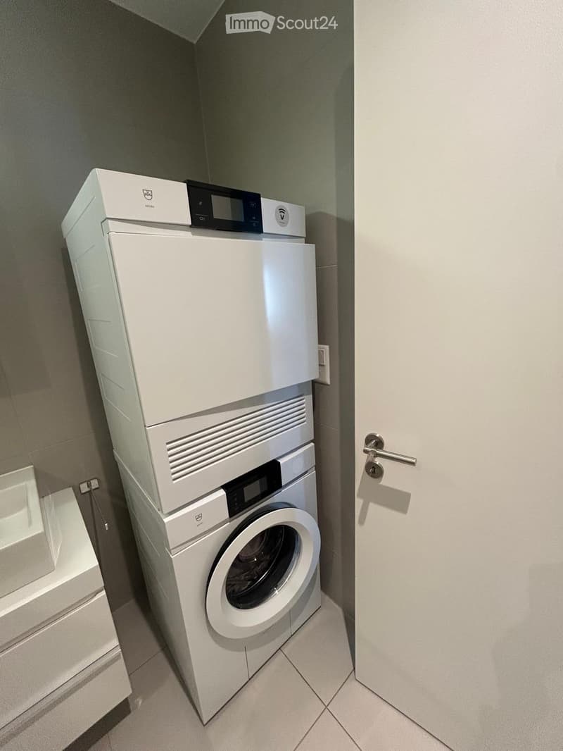 A small room with a white washing machine and dryer stacked on top of each other. A white sink and cabinet are on the left side. The room has white tiled flooring and walls.