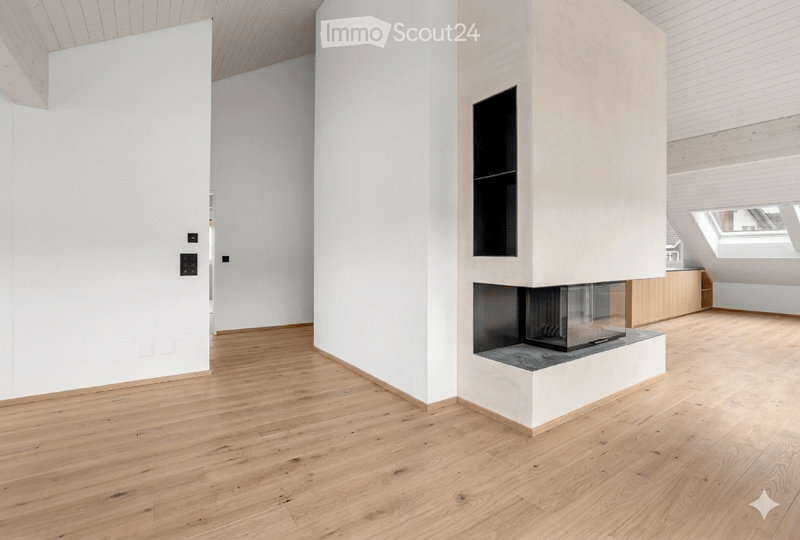 Bright, minimalist room with light wooden flooring, white walls, and a modern fireplace with a glass front. The ceiling features exposed wooden beams and skylights, allowing natural light to fill the space.