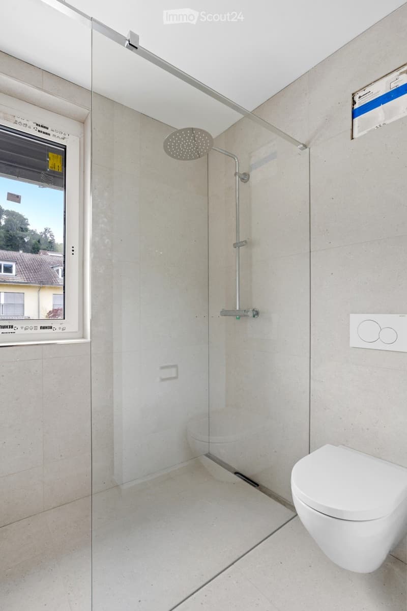 Modern bathroom with a glass shower enclosure, a white toilet, and a sleek shower head mounted on the wall. The space features light-colored tiles and a large window offering a view of the outside.