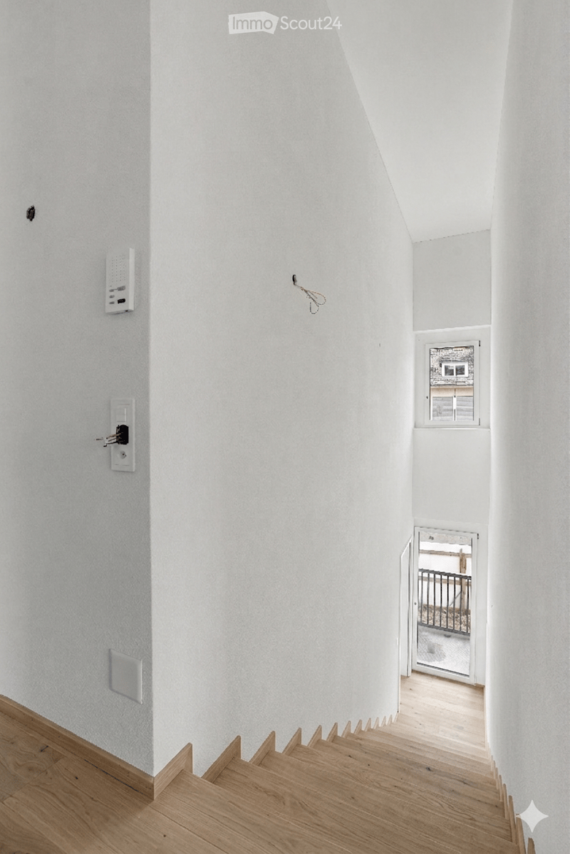 A narrow, modern hallway with white walls, wooden flooring, and a small window at the end. The hallway features a small staircase leading upwards, with a door opening to an outdoor area.