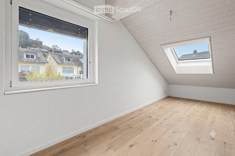 Empty attic room with wooden flooring, white walls, and large windows offering a view of neighboring houses.