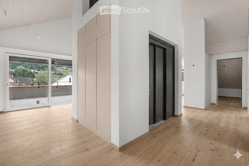 Bright, open hallway with wooden flooring, white walls, large glass doors leading to a balcony with scenic countryside view.