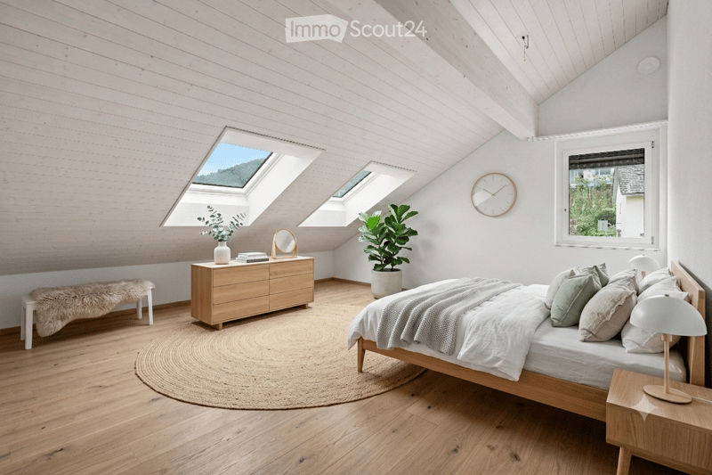 Bright, modern bedroom with wooden floors, white walls, and skylights. Features a wooden bed with white bedding, a nightstand with a lamp, a dresser with a mirror, a bench, a round rug, and potted plants.