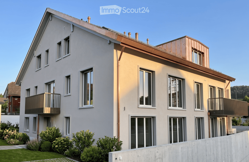 Modern two-story house with white exterior, multiple windows, and balconies. Features a sloped roof with copper accents and a well-maintained garden in front.