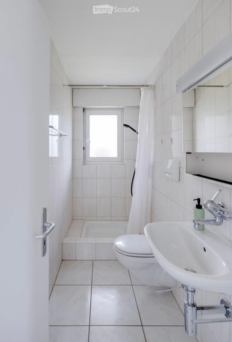 Bathroom with white tiles, toilet, shower, sink, soap dispenser, and mirror.
