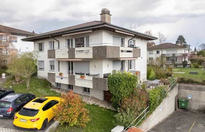 Three-story apartment building with balconies, cars parked in front, and a garden area.
