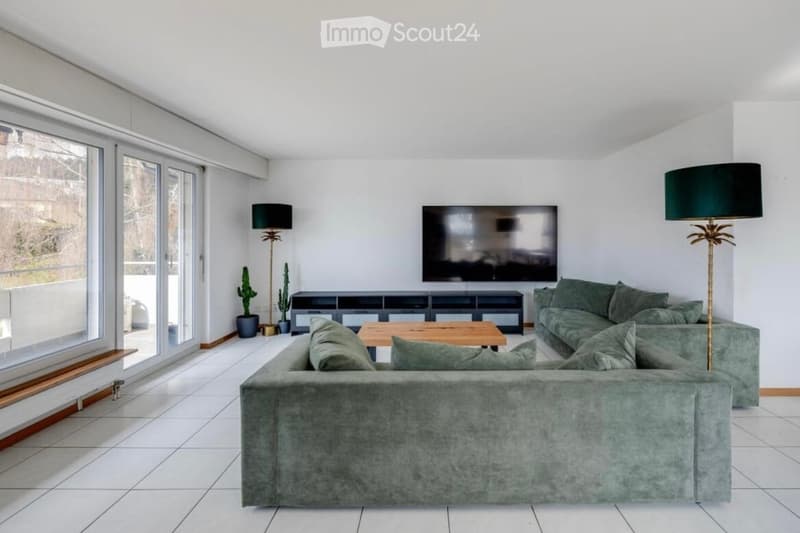 Modern living room with white tiles, large windows, and a balcony. Features a large green couch, a wooden coffee table, floor lamps, and a flat-screen TV.