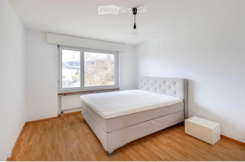 A room with a bed, a nightstand, and a lamp. The room has white walls and wooden floors.