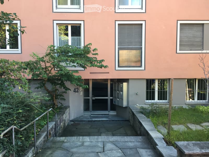 A three-story building with a pink facade, white windows, and a gray entrance with a mailbox. The entrance is flanked by a tree and a small garden. The building has a number 42 on the wall above the entrance.