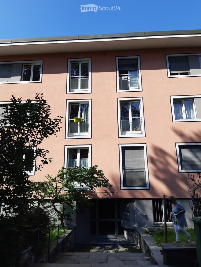 A pink building with multiple windows and a balcony on the second floor. A woman is walking towards the entrance.