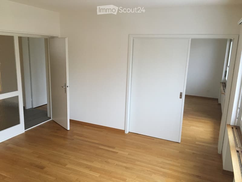 An empty room with white walls and wooden floors. There are two doors, one of which is open, leading to another room. The room has a glass window on the right side. The floor is clean and well-maintained.